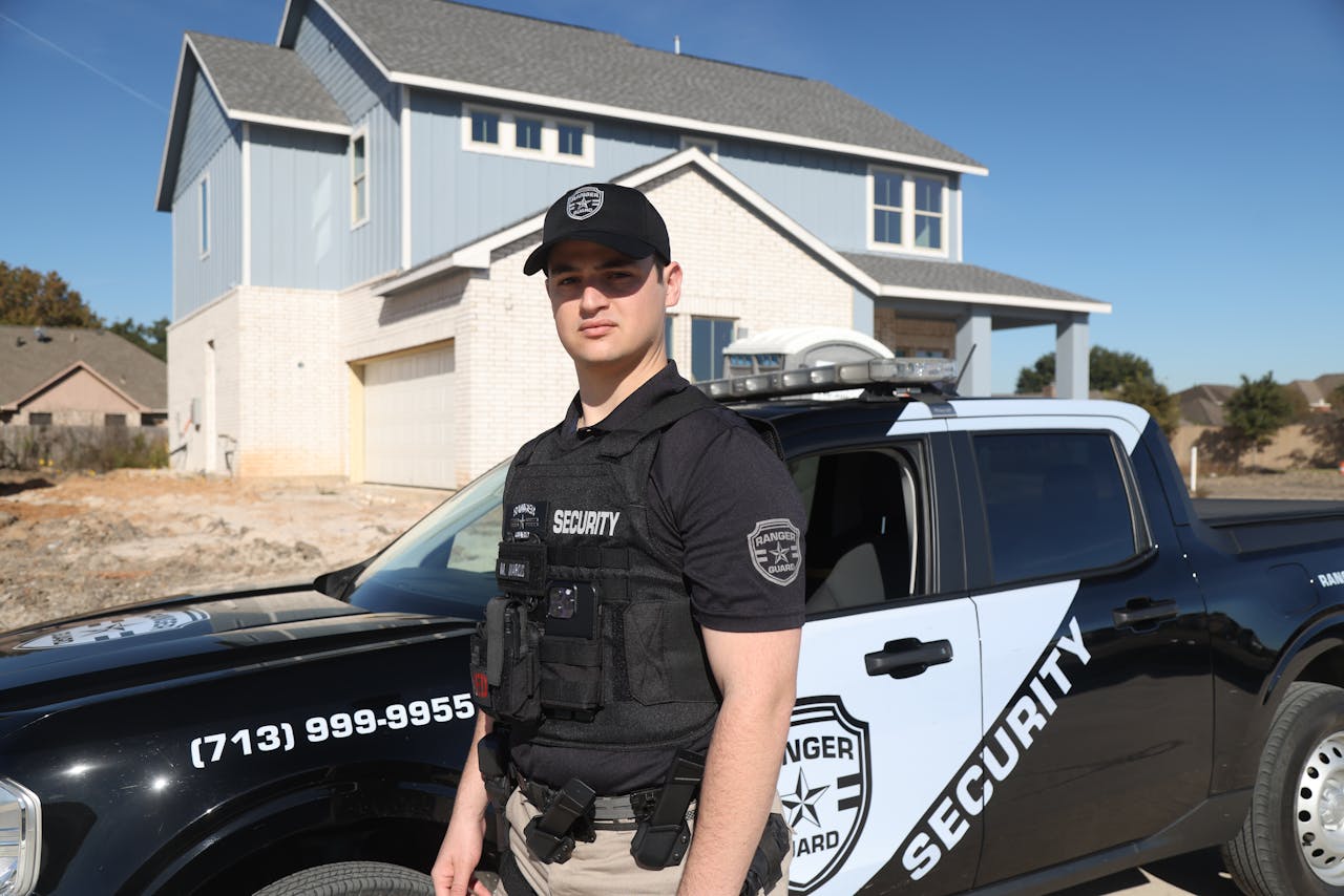 Security guard in uniform stands beside patrol car outside residential building in daylight.